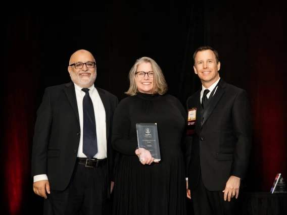 Two men in suits stand on each side of a woman in a black dress holding a glass award. 