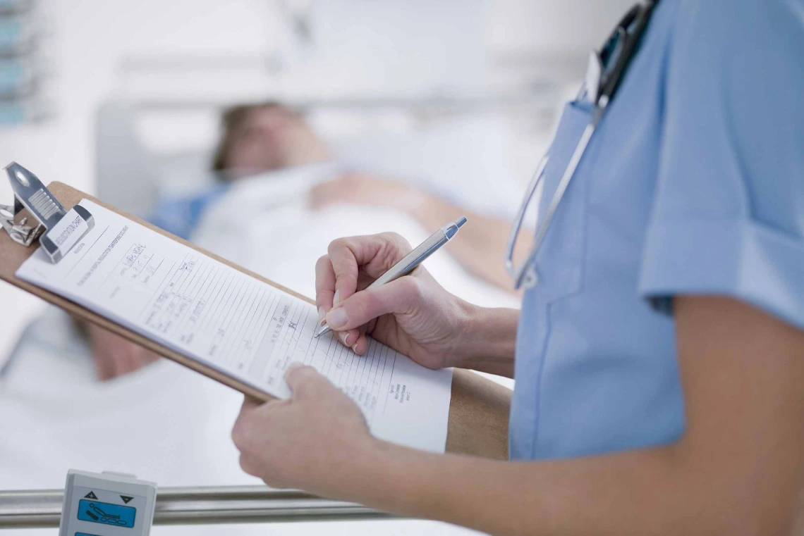 Close-up of nurse in light blue scrubs with stethoscope around neck writing on a clipboard with a hospital patient blurred in the background