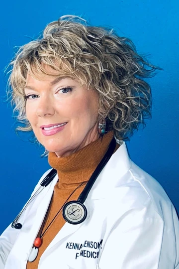 Woman doctor with short, curly blonde hair stands against a blue background, wearing a burnt orange turtleneck, white coat with her name on it and a stethoscope.