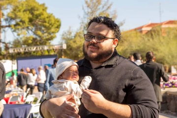 A man holds a baby tight to his chest as he stands in the shade of a tree outdoors.