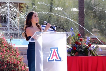 A young woman speaks into a microphone while standing behind a lectern bearing the University of Arizona logo. A fountain is in the background.
