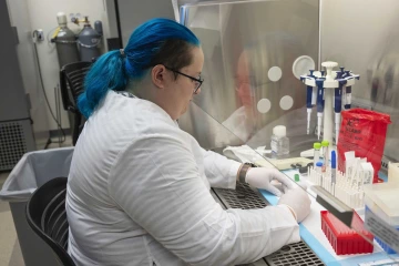A person wearing a white lab coat works at a laboratory bench, handling scientific equipment and processing samples.