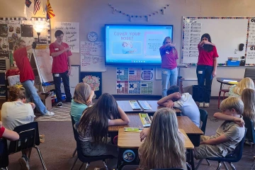 College students in red shirts stand in front of a class and demonstrate how to cover your nose as part of a health presentation