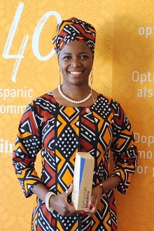 Dark-skinned woman wearing a head wrap and patterened dress stands in front of a 40 Under 40 banner holding an award and smiling.
